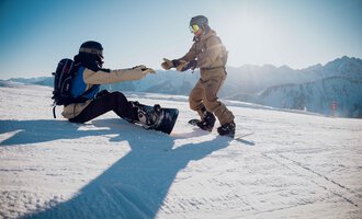 Zwei Snowboarder auf einer verschneiten Piste. Einer hilft dem anderen, der auf dem Boden sitzt. | © Skischule Jörihalde Kleinwalsertal | Ole Zumpolle
