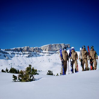 Eine Gruppe von Skifahrern steht auf einer schneebedeckten Fläche. Im Hintergrund sieht man beeindruckende Berggipfel und einen klaren blauen Himmel. | © Skischule Jörihalde Kleinwalsertal | Herbert Jochum