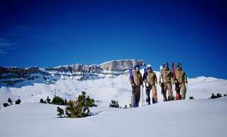 Eine Gruppe von Skifahrern steht auf einer schneebedeckten Fläche. Im Hintergrund sieht man beeindruckende Berggipfel und einen klaren blauen Himmel. | © Skischule Jörihalde Kleinwalsertal | Herbert Jochum