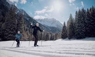 Zwei Personen beim Langlaufen in einer verschneiten Landschaft. Im Hintergrund sind majestätische Berge und eine klare, sonnige Himmel zu sehen. | © Skischule Jörihalde Kleinwalsertal | Ole Zumpolle