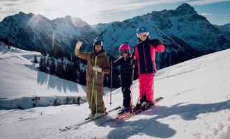 Drei Skifahrer stehen auf einem verschneiten Hang und winken in die Kamera. Im Hintergrund sind beeindruckende Berge und ein klarer Himmel zu sehen. | © Skischule Jörihalde Kleinwalsertal | Ole Zumpolle