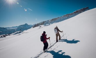 Zwei Wanderer gehen durch tiefen Schnee in den Bergen. Die Sonne scheint klar am blauen Himmel. | © Skischule Jörihalde Kleinwalsertal | Ole Zumpolle