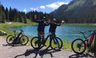 Zwei Radfahrer stehen am Ufer eines klaren Sees und lächeln fröhlich in die Kamera. Im Hintergrund sind grüne Berge und ein klarer blauer Himmel zu sehen. | © WalserBike Tours | Christian Gutermuth