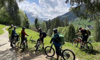 Eine Gruppe von Radfahrern steht auf einem Weg in den Bergen. Die Umgebung ist grün mit Wald und einer beeindruckenden Berglandschaft im Hintergrund. | © WalserBike Tours | Christian Gutermuth