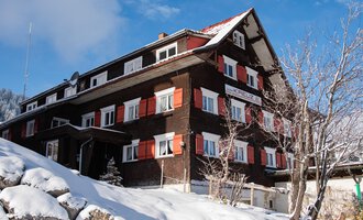 Ein großes, traditionelles Chalet in den Bergen, umgeben von Schnee. Die Fenster sind mit roten Fensterläden dekoriert, und der Himmel ist klar und blau.
