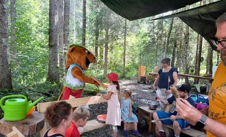 Ein niedlicher Maskottchen begrüßt Kinder im Wald. Einige Erwachsene und Kinder sitzen an einem Tisch und genießen die Zeit in der Natur. | © Wildnisschule Kleinwalsertal