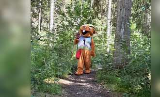 Ein Maskottchen in einem Wald trägt eine Trikotnummer. Es steht auf einem schmalen Pfad zwischen den Bäumen. | © Wildnisschule Kleinwalsertal