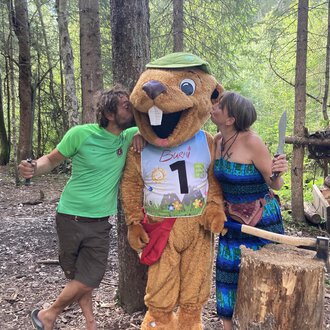 Ein lustiger Maskottchen in Form eines Bibern steht zwischen zwei Personen im Wald. Die Menschen scheinen Spaß zu haben und küssen das Maskottchen auf die Wangen. | © Wildnisschule Kleinwalsertal
