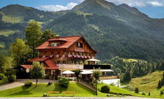 Ein malerisches Chalet in den Bergen mit blühenden Fensterkästen. Im Hintergrund erheben sich grüne Hügel und majestätische Berge.