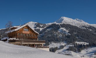 A rustic chalet in the snow, surrounded by majestic mountains. The clear blue sky makes the winter landscape particularly shine. | © Wirtshaus Hoheneck | Klaus Ruland