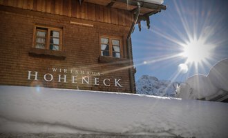 A cozy wooden house with the inscription "Wirtshaus Hoheneck" under a blanket of snow. The sun shines bright over the snowy surroundings.