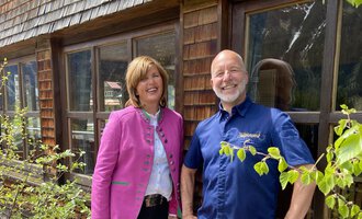 A man and a woman are standing smiling in front of a wooden building. The woman is wearing a pink jacket and the man a blue shirt. | © Wirtshaus Hoheneck | Klaus Ruland
