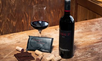 A bottle of red wine with a wine glass, chocolate, and cork on a wooden table. The wine is presented in an elegant packaging. | © Wirtshaus Hoheneck | Klaus Ruland