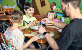 Ein Junge isst mit Freude Pizza, während er mit zwei anderen Kindern an einem Tisch sitzt. Im Hintergrund sind bunte Wandmalereien zu sehen. | © Wonniland Sonthofen | interSPA Gruppe