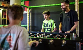 Zwei Jungen spielen Tischfußball, während ein Erwachsener zuschaut. Die Atmosphäre ist lebhaft und freundlich. | © Wonniland Sonthofen | interSPA Gruppe
