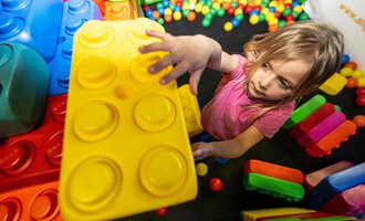 Ein kleines Kind spielt mit großen, bunten Bausteinen in einem Spielbereich. Es hält einen gelben Baustein in der Hand und scheint konzentriert zu sein. | © Wonniland Sonthofen | interSPA Gruppe