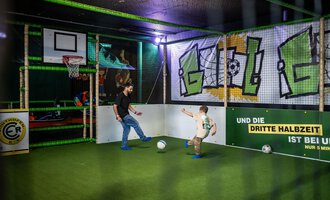 Ein spielender Vater und sein Kind in einer aufregenden Indoor-Sportarena. Sie kicken mit einem Fußball auf grünem Kunstrasen unter bunten Grafiken. | © Wonniland Sonthofen | interSPA Gruppe
