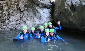 Eine Gruppe von acht Personen in Neoprenanzügen und grünen Helmen steht im Wasser zwischen großen Felsen. Sie lächeln und zeigen Daumen hoch. | © Rocksolid Canyoning Tours