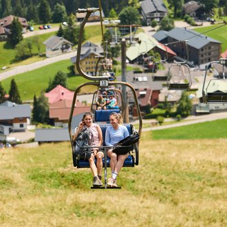 Two women are sitting in a chair lift and riding up a mountain meadow. In the background, green hills and a small village with houses can be seen. | © Kleinwalsertal Tourismus | Oliver Farys