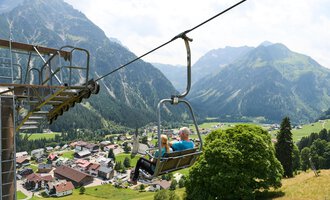 A gondola lifts over a picturesque valley with mountains in the background. In the foreground, there is a quaint place with red roofs and green meadows. | © Kleinwalsertal Tourismus | Oliver Farys