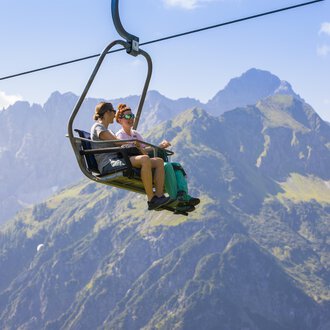 Two people are sitting on the chairlift and enjoying the view of the mountains. In the background, green meadows and high peaks can be seen. | © Kleinwalsertal Tourismus | Frank Drechsel