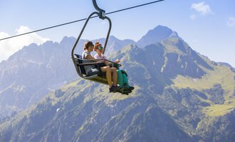 Two people are sitting on the chairlift and enjoying the view of the mountains. In the background, green meadows and high peaks can be seen. | © Kleinwalsertal Tourismus | Frank Drechsel