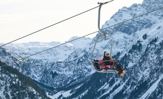 A person is riding in a chairlift over snow-covered mountains. The landscape is majestic and showcases the wintry beauty of the Alps. | © Kleinwalsertal Tourismus | Oliver Farys