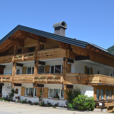 Ein traditionelles alpenländisches Haus mit Holzverkleidung und mehreren Balkonen. Im Hintergrund sind grüne Berge und ein blauer Himmel zu sehen.