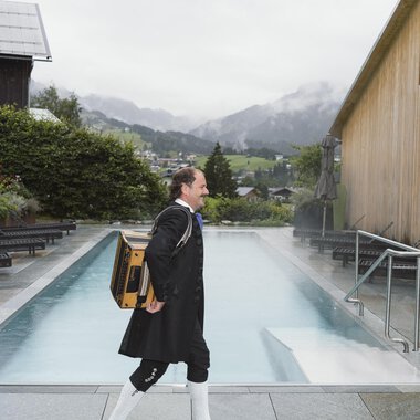 Ein Mann in traditioneller Tracht trägt eine Kiste und geht an einem Pool entlang. Im Hintergrund sind Berge und Nebel zu sehen.