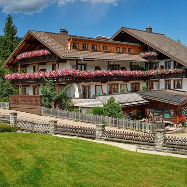 Ein schönes alpenländisches Hotel mit bunten Blumen auf dem Balkon. Umgeben von grünen Wiesen und Bäumen unter einem blauen Himmel.