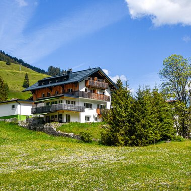 Ein malerisches Haus inmitten einer grünen Wiese und umgeben von Bäumen. Im Hintergrund sind sanfte Hügel unter einem klaren blauen Himmel zu sehen.