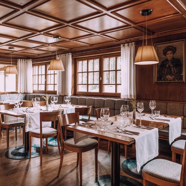 Ein gemütliches Restaurant mit Holzwänden und eleganten Tischdeckern. Die Tische sind gedeckt und es gibt große Fenster, die viel Licht hereinlassen.