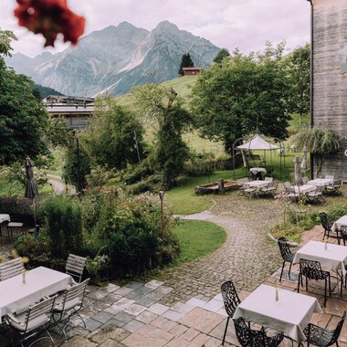 Ein einladender Außenbereich eines Restaurants mit Tischen und Stühlen. Im Hintergrund sind grüne Bäume und majestätische Berge zu sehen.