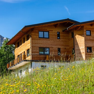 Ein modernes Holzhaus steht in einer grünen Wiese, umgeben von Bäumen und Bergen. Der Himmel ist klar und blau.