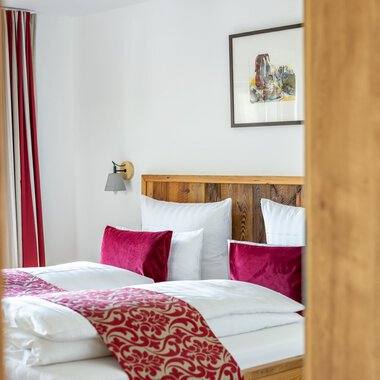 Ein gemütliches Schlafzimmer mit einem großen Bett und roten Kissen. Die Wände sind hell und es gibt ein Kunstwerk im Hintergrund.