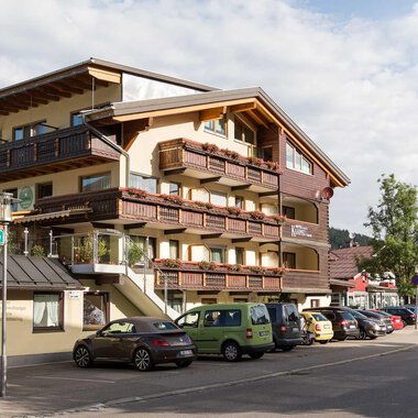 Ein modernes Gebäude mit mehreren Balkonen liegt an einer Straße. Umgeben von Bäumen und Parkplätzen strahlt es eine einladende Atmosphäre aus.