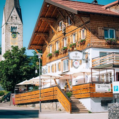 Ein traditionelles Holzgebäude mit einer einladenden Terrasse und einem schönen begrünten Bereich. Im Hintergrund ist ein Kirchturm visible.