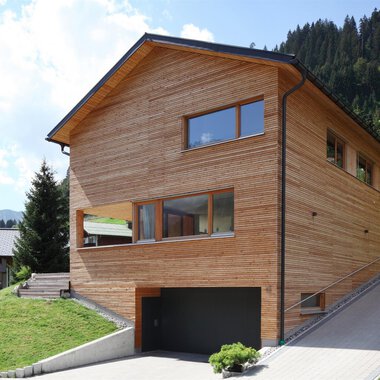 Ein modernes Holzhaus in einer grünen Umgebung mit Bergen im Hintergrund. Die Architektur ist schlicht und harmoniert gut mit der Natur.