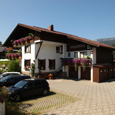 Ein charmantes Gebäude im bayerischen Stil mit bunten Blumen an den Fenstern. Der Parkplatz vor dem Haus ist gut beleuchtet und bietet Platz für mehrere Autos.
