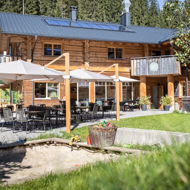 Ein moderner Hof umgeben von grüner Natur. Auf der Terrasse stehen Tische und Stühle mit Sonnenschirmen für entspannte Stunden im Freien.