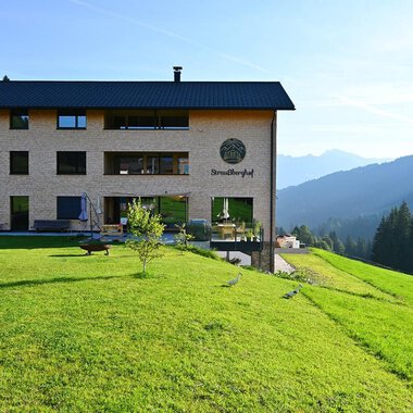 Ein modernes Haus in den Bergen, umgeben von grünen Wiesen und Wald. Im Hintergrund sind sanfte Hügel und ein klarer Himmel zu sehen.