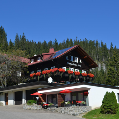 A pretty building with a balcony full of flower boxes and red umbrellas. In the background, there are green trees and a clear sky.