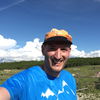 Ein lachender Mann in blauer T-Shirt und orange Mütze steht in einer grünen Landschaft bei schönem Himmel.