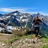 Frau beim Wandern in den Bergen mit Schnee auf den Gipfeln, trägt Rucksack und Wanderstöcke, sonniger Tag.