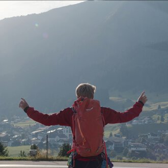 Eine Person mit rotem Rucksack steht auf einer Wiese und zeigt nach oben, während ein Hubschrauber im Flug über der Landschaft fliegt.