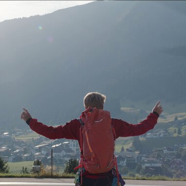 Eine Person mit rotem Rucksack steht auf einer Wiese und zeigt nach oben, während ein Hubschrauber im Flug über der Landschaft fliegt.