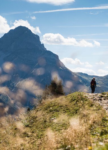 Ein Wanderer geht auf einem Pfad in den Bergen im Schwarzwald bei schönem Wetter, umgeben von grünem Gras und Bergen im Hintergrund. | © Kleinwalsertal Tourismus eGen • Fotograf: Andre Tappe