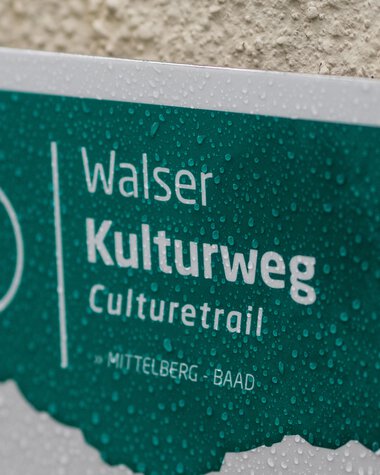 Hinweisschild für den Kulturweg Walters in Mittelberg-Baad, feucht vom Regen, mit Wassertröpfchen auf der Oberfläche. | © Kleinwalsertal Tourismus eGen • Fotograf: Andre Tappe