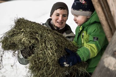 Eine Frau und ein Kind, die im Schnee beim Wildfütterung im Wald stehen, halten eine große Portion Heu oder Wildfutter zusammen. | © Kleinwalsertal Tourismus eGen • Fotograf: Andre Tappe