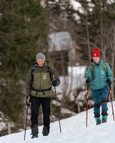 Zwei Menschen wandern in winterlicher Natur durch den Schnee, beide mit Rucksack und Wanderstöcken. | © Kleinwalsertal Tourismus eGen • Fotograf: Basti Heckl